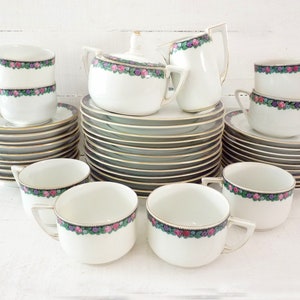 Könnte beinhalten: Ein Vintage-Teeservice aus weißem Porzellan mit Goldrand. Das Set umfasst Tassen, Untertassen, Teller, eine Zuckerdose und einen Sahnekännchen. Jedes Stück ist mit einem Blumenband aus rosa, lila und grünen Blumen verziert. Die Teller und Untertassen haben einen dunkelgrünen Rand.