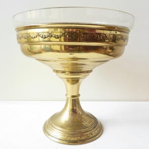 Può includere: Una ciotola in metallo dorato con un inserto in vetro poggia su una base a piedistallo. La ciotola ha un bordo decorativo e la base ha un design scanalato.