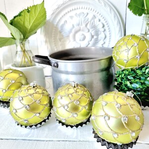 Pode incluir: Cinco bolas de Natal verdes com detalhes dourados e prateados. As bolas são redondas e têm um acabamento brilhante. Estão sobre uma superfície branca.