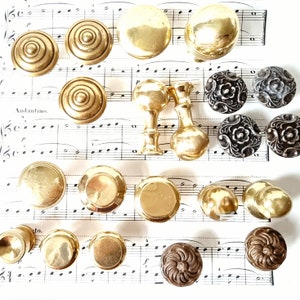 May include: Assortment of decorative cabinet knobs in gold, silver, and black finishes. Some knobs have a floral design, while others have a simple round shape. The knobs are arranged on a sheet of music.