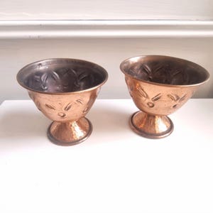 May include: Two copper-coloured metal goblets with a hammered texture and decorative floral patterns. The goblets have a wide bowl shape and are set on a pedestal base. The interior of the goblets is a darker shade of copper.