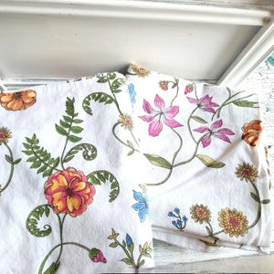 May include: White floral fabric featuring a repeating pattern of colorful flowers and green foliage. The flowers are in shades of pink, orange, yellow, and blue. The fabric is folded and appears to be a tablecloth or similar textile.