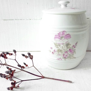 May include: White ceramic jar with a lid, featuring a floral design with pink and grey flowers. The jar is sitting on a white surface.