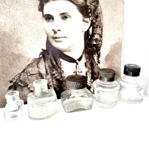 Könnte beinhalten: Fünf antike Glas-Tintenflaschen in verschiedenen Formen und Größen. Die Flaschen haben braune, schwarze und klare Glasverschlüsse. Die Flaschen sind auf einer weißen Oberfläche angeordnet.