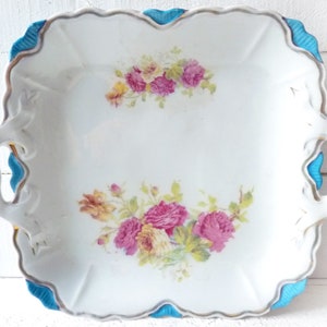 May include: A white porcelain serving dish with blue and gold trim. The dish has two handles and features a floral design with pink and yellow roses.