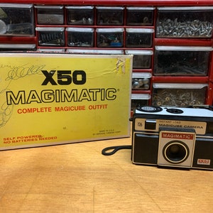 May include: A vintage "Magimatic" instant load magicube camera with a black and silver finish, and a matching box. The box is yellow with the text "X50 MAGIMATIC COMPLETE MAGICUBE OUTFIT". The camera is labeled "126" and "Instant Load Magicube Camera".