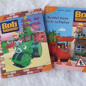 Könnte beinhalten: Zwei Bob der Baumeister Kinderbücher. Das erste Buch heißt "Bob der Baumeister Rollo, der Tollpatsch" und zeigt ein grünes und gelbes Baufahrzeug. Das zweite Buch heißt "Bob der Baumeister Buddel kann nicht schlafen" und zeigt ein rotes Baufahrzeug.