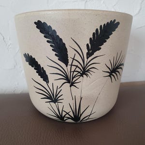 Könnte beinhalten: Ein gesprenkelter beigefarbener Keramikübertopf mit einem handgemalten schwarzen botanischen Motiv. Das Design zeigt mehrere Weizen- oder Grasähren mit dünnen, spitzen Blättern und strukturierten, federartigen Spitzen auf der Vorderseite.