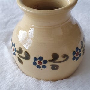 Könnte beinhalten: Eine kleine, cremefarbene Keramikvase mit rundem Körper und einer breiten Öffnung. Die Vase ist mit einem floralen Muster in Blau-, Rot- und Grüntönen verziert.