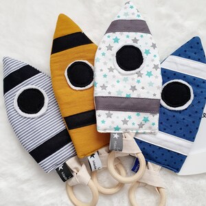 Könnte beinhalten: Vier handgefertigte Baby-Spielzeuge in Raketenform. Die Spielzeuge sind in verschiedenen Mustern gestaltet: gestreift, senfgelb mit schwarzen Streifen, Sterne auf weißem Grund und blaue Punkte. Jedes Spielzeug hat einen Holzring zum Greifen.