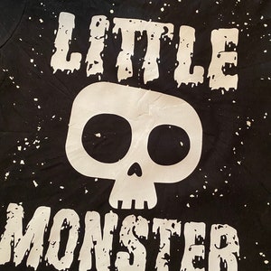 May include: Black t-shirt with white splatter paint effect and the words "LITTLE MONSTER" in bold, white letters. A white skull graphic is centered between the words. The shirt is likely for children.