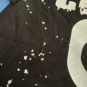 May include: Close-up of a black t-shirt with white lettering and light green paint splatters. The fabric is textured, and the design appears to be screen-printed. The shirt has a casual, artistic aesthetic.