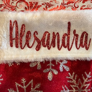 May include: A red Christmas stocking with the name "Alesandra" in glittery red script on a white faux fur cuff. The stocking has a red velvet body with silver snowflake designs.