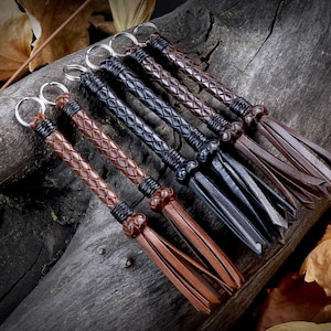 May include: A collection of leather keychains with braided handles and tassel ends. The keychains are in various shades of brown and black, with silver key rings. The items are arranged on a dark wooden surface with autumn leaves.