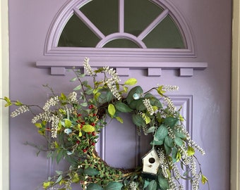 May include: A wreath made of greenery, white flowers, and red berries hangs on a lavender door. A small birdhouse is attached to the wreath.