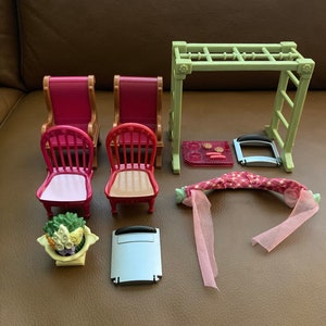 May include: A set of dollhouse furniture including two red chairs, a green jungle gym, a pink and white blanket, a white and gray stovetop, and a white planter with green and yellow flowers.