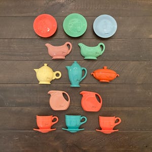 May include: A collection of colorful ceramic dishware in various shapes and sizes. The set includes plates, teapots, pitchers, gravy boats, and cups in shades of red, green, blue, yellow, teal, and orange. The dishware is arranged on a dark wooden surface.