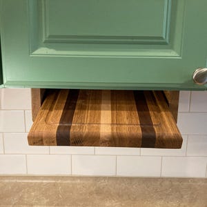 May include: A wooden cutting board with a light brown, dark brown, and black striped design is mounted under a green cabinet. The cutting board is pulled out and ready to use.