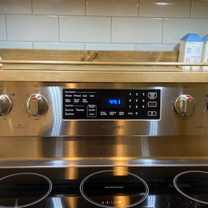 May include: A stainless steel Samsung electric range with a wooden shelf. The shelf holds a plush toy and two decorative house-shaped salt and pepper shakers. The range has four burners and a digital display showing the time.