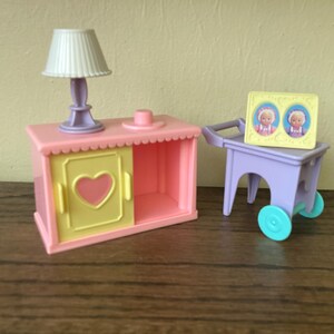 May include: A pink and yellow toy cabinet with a heart-shaped cutout on the door. A purple toy cart with two blue wheels and a picture frame with two dolls inside. The picture frame has a yellow background and the dolls are wearing pink and white dresses.