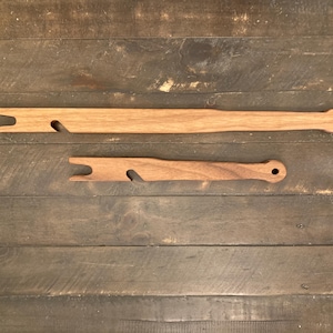 May include: Two wooden weaving tools, a shuttle and a pick-up stick, rest on a wooden surface. The shuttle has a forked end and a curved cutout. The pick-up stick is a flat, rectangular piece with a hole at one end.