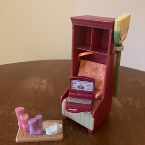May include: A miniature dollhouse furniture set featuring a red armchair with a laptop, a shelf unit, and a small tray with shoes and boots. A green and yellow brush is attached to the side of the shelf unit. The set is made of plastic.