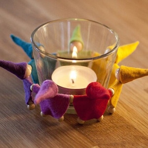 Könnte beinhalten: Ein Glas-Kerzenhalter mit einem Teelicht, umgeben von fünf Filzfiguren in einem Kreis. Die Figuren sind in verschiedenen Farben: lila, rot, gelb, blau und grün.