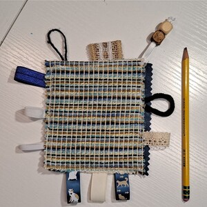 May include: A square sensory toy with a woven blue, tan, and white pattern, featuring various textures and attachments. Includes ribbons, lace, and a loop, with a pencil for size reference. The ribbons have cat illustrations.