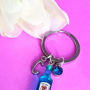 May include: A blue glass bottle keychain with a silver charm that says "C" and a blue crystal charm. The bottle has a label that says "Fast King Dry Gin".