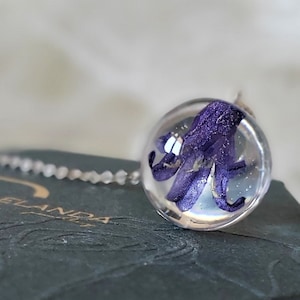 May include: A purple flower encased in a clear glass sphere pendant on a silver chain. The pendant is sitting on a dark green box with a textured surface.