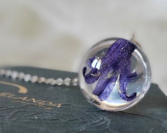 Puede incluir: Un colgante de flor morada encerrada en una esfera de vidrio transparente en una cadena de plata. El colgante está sobre una caja verde oscuro con una superficie texturizada.