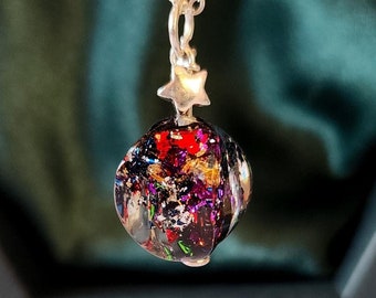 May include: A silver chain necklace with a star charm and a round, multi-colored glass pendant. The pendant is filled with iridescent glitter and has a black background.