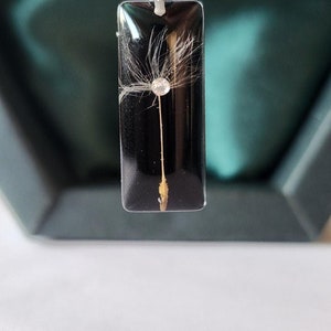 May include: A rectangular pendant necklace with a silver chain. The pendant is black with a dandelion seed head encased within. A small, clear crystal is set in the center of the seed head.