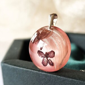 May include: A spherical, pink resin pendant with a silver bail. The pendant features a dried flower and other organic elements. The pendant is presented in a dark green jewellery box, with a soft, blurred background.