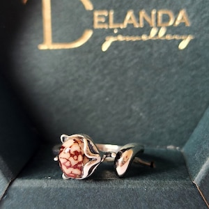 Puede incluir: Un anillo de plata con una piedra marrón y blanca engastada en un diseño floral. El anillo está en una caja verde con el texto "Delanda Jewelry" en la tapa.
