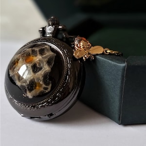 Peut inclure: Une montre de poche noire avec un cadran transparent révélant un intérieur à motifs. Un charm en forme d'abeille de couleur or est suspendu à la montre. La montre est posée sur une boîte vert foncé.