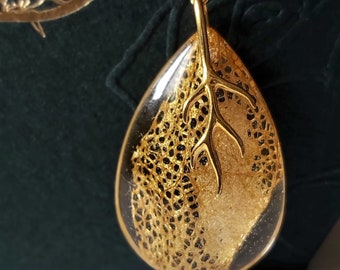 May include: A gold necklace with a teardrop-shaped pendant featuring a delicate gold leaf design and a small gold branch. The pendant is set against a black background.