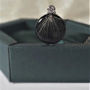 Puede incluir: Un colgante circular negro con un cristal transparente en la parte superior. El colgante tiene un dise&ntilde;o delicado e intrincado de l&iacute;neas finas que irradian desde el centro.