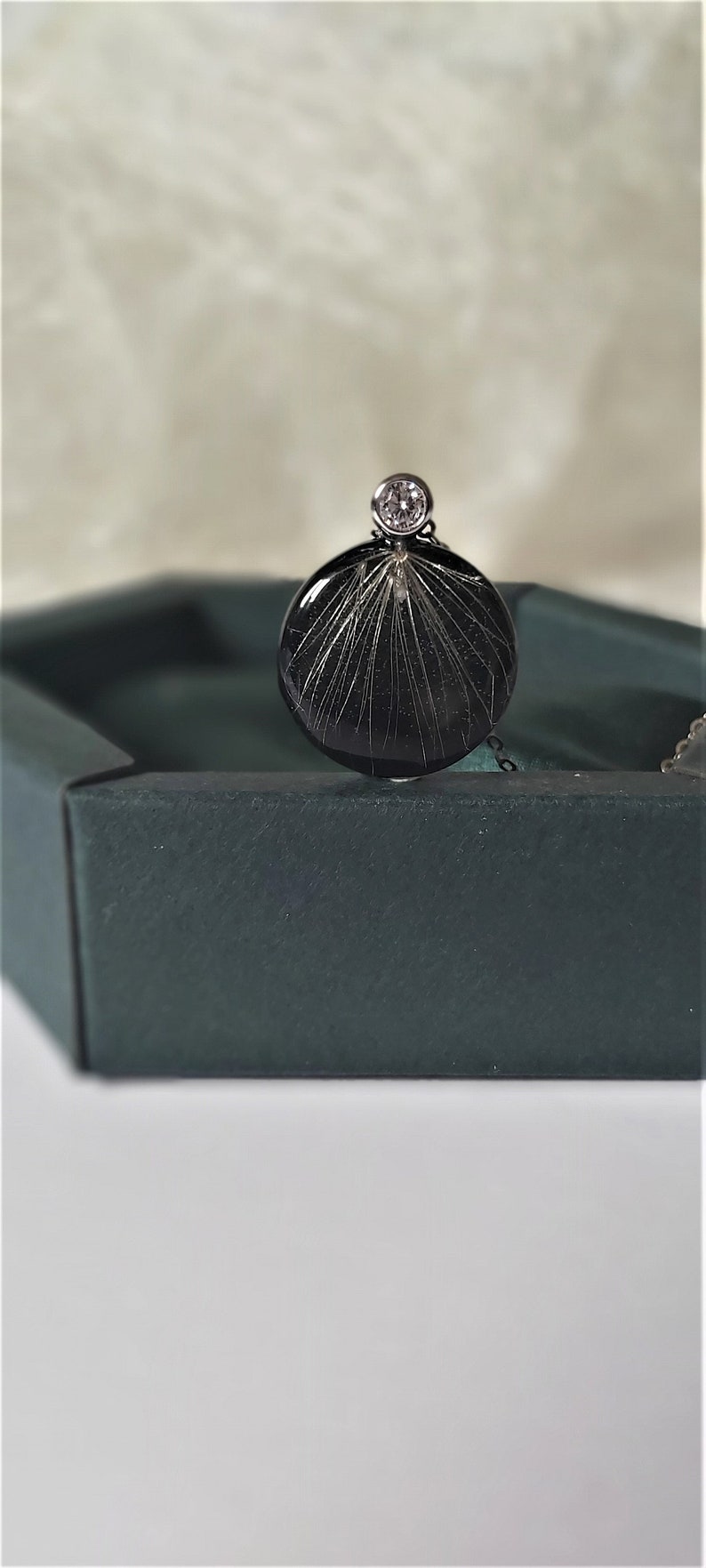 Puede incluir: Un colgante circular negro con un cristal transparente en la parte superior. El colgante tiene un dise&ntilde;o delicado e intrincado de l&iacute;neas finas que irradian desde el centro.