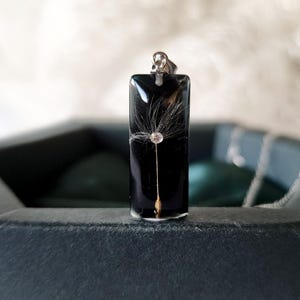 May include: A rectangular black pendant with a dandelion seed head encased in clear resin. The pendant has a silver-colored bail and is displayed in a dark gray box. The dandelion seed head is white and delicate.