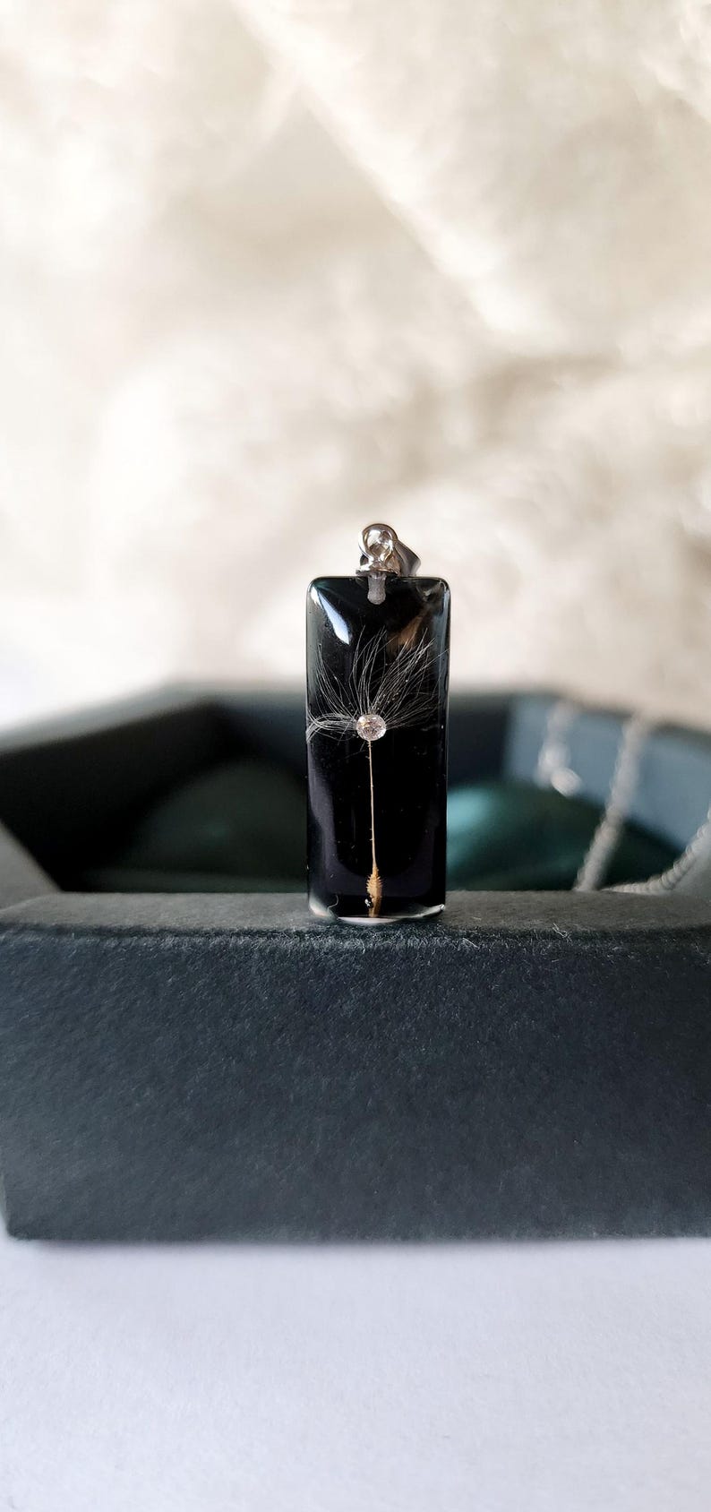 May include: A rectangular black pendant with a dandelion seed head encased in clear resin. The pendant has a silver-colored bail and is displayed in a dark gray box. The dandelion seed head is white and delicate.