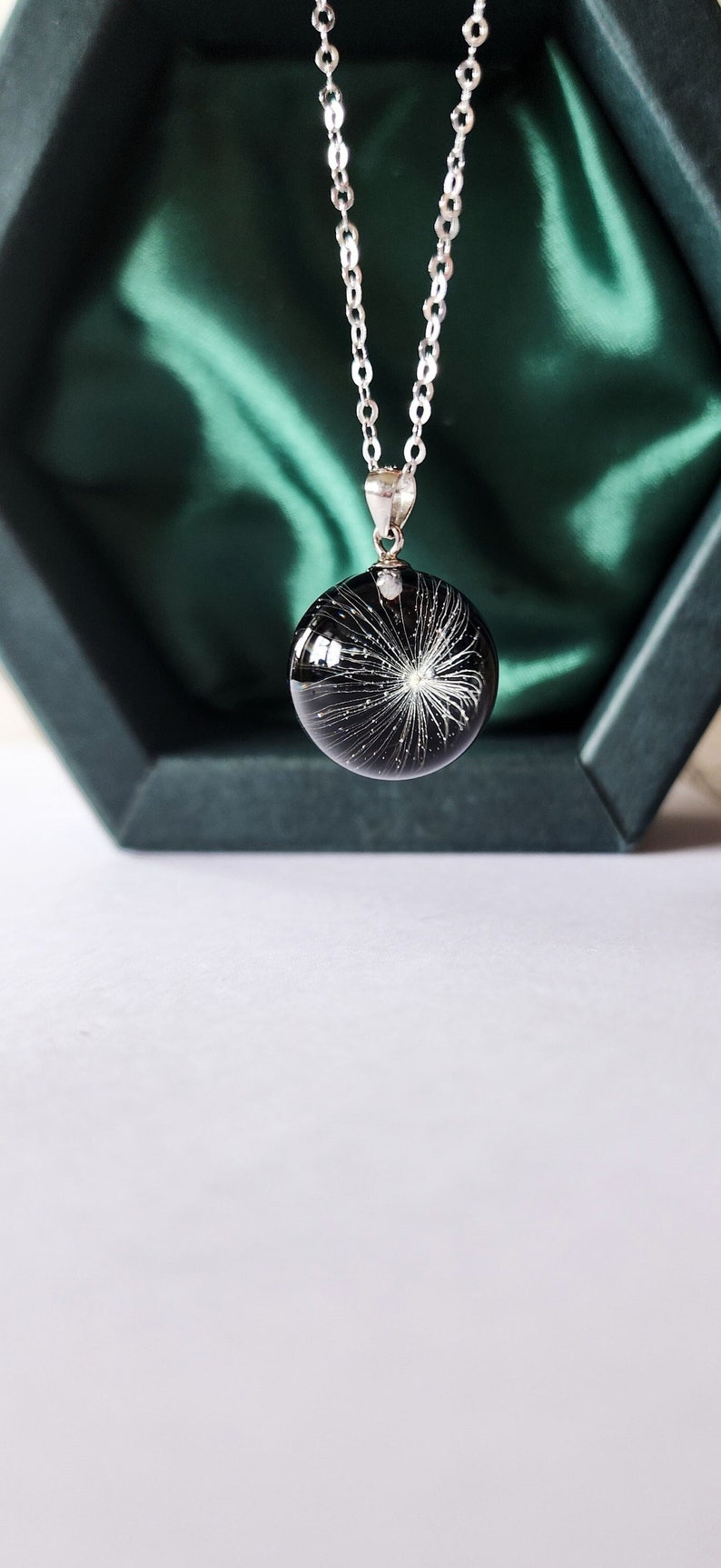 May include: A silver necklace with a round, black pendant. The pendant features a white, starburst-like design. The necklace is displayed against a dark green background, creating a striking contrast. The pendant is approximately 1 inch in diameter.