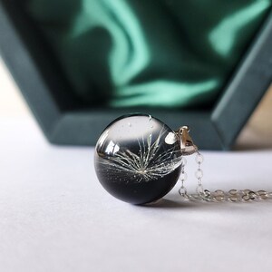 May include: A clear, spherical pendant necklace with a silver chain. The pendant contains a black and white botanical design. The necklace is displayed on a white surface, with a dark green box in the background.