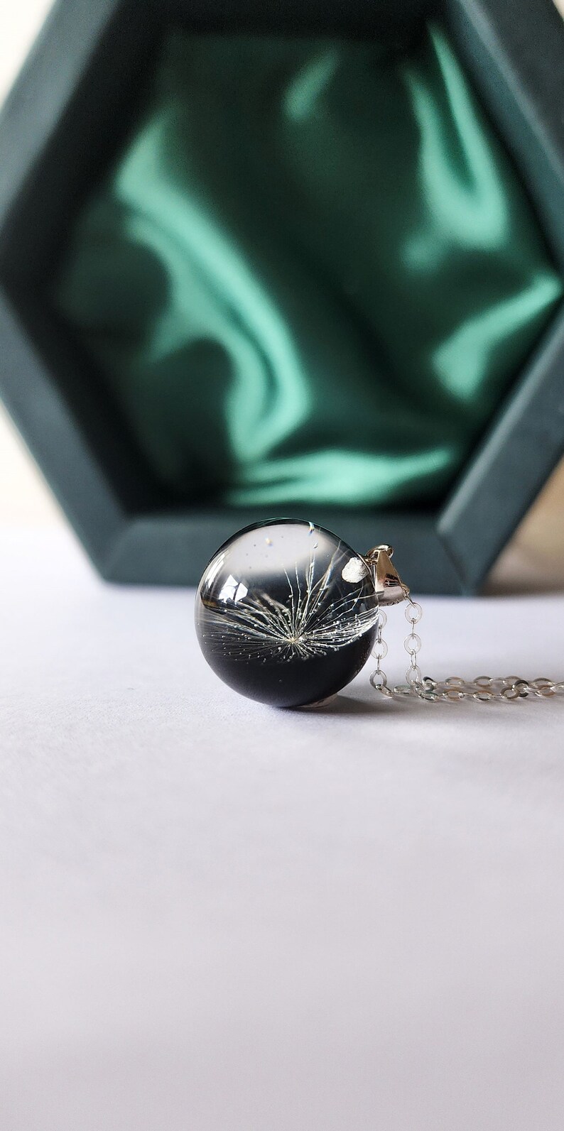 May include: A clear, spherical pendant necklace with a silver chain. The pendant contains a black and white botanical design. The necklace is displayed on a white surface, with a dark green box in the background.