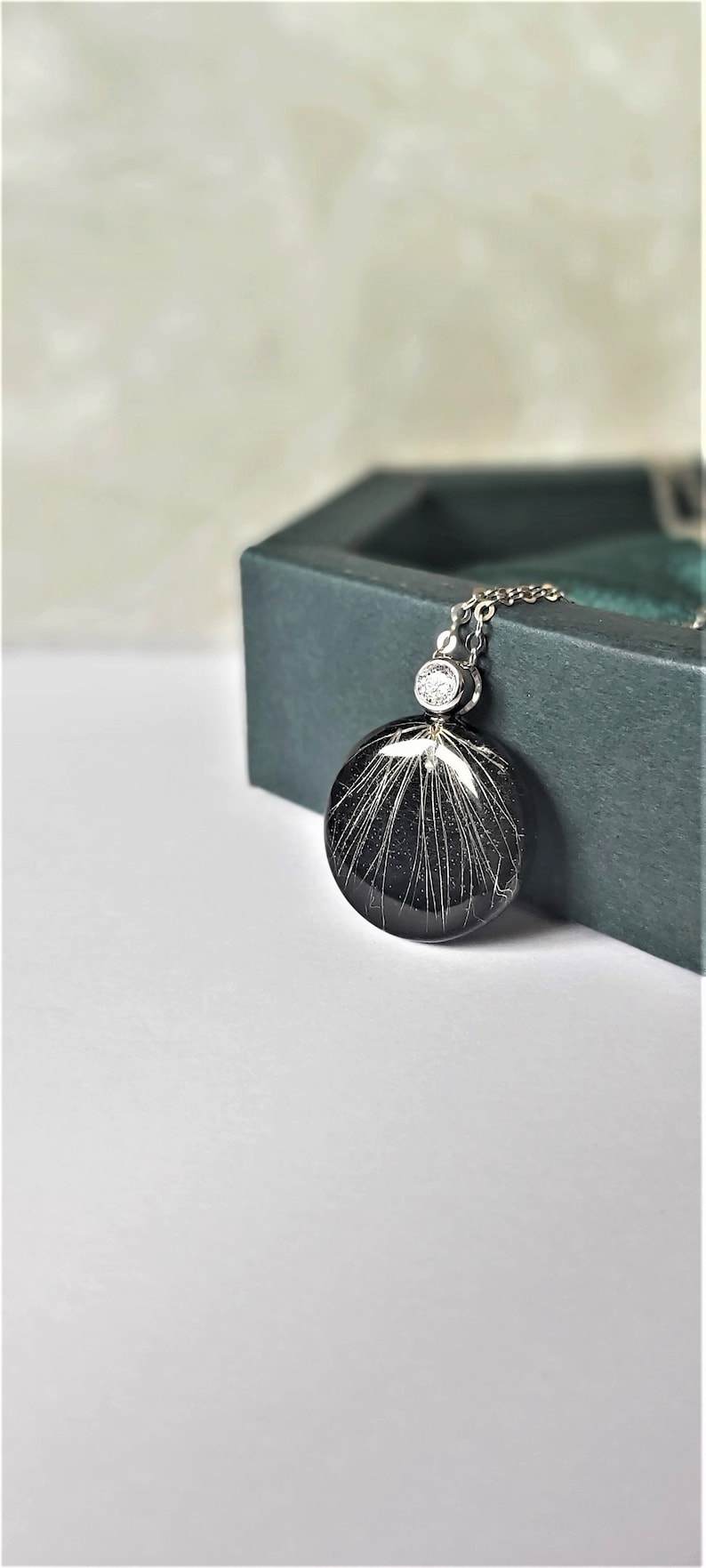 Puede incluir: Un collar de cadena de plata con un colgante negro que presenta un dise&ntilde;o blanco brillante. El colgante es redondo y tiene un peque&ntilde;o cristal transparente en la parte superior.