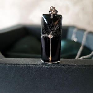 May include: A rectangular black pendant with a dandelion seed encased in clear resin. The pendant has a silver bail and is displayed in a dark gray box. The dandelion seed is centered in the pendant.