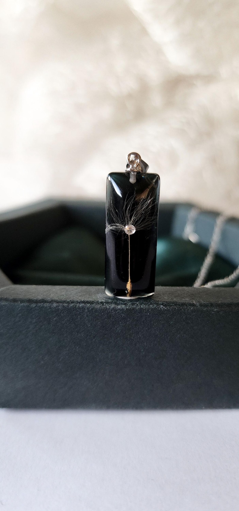 May include: A rectangular black pendant with a dandelion seed encased in clear resin. The pendant has a silver bail and is displayed in a dark gray box. The dandelion seed is centered in the pendant.
