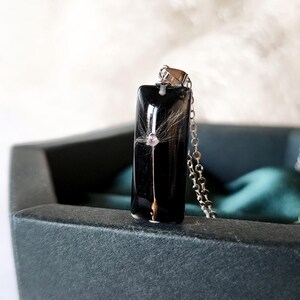 May include: A rectangular black pendant necklace with a dandelion seed encased in clear resin. The pendant hangs from a silver chain. The necklace is displayed in a dark green box.