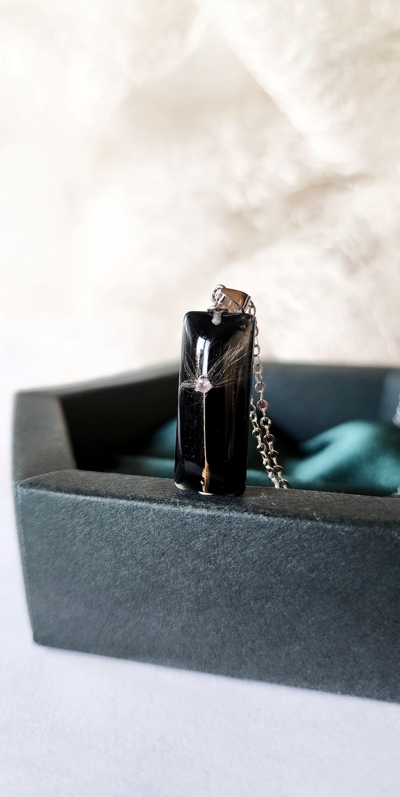 May include: A rectangular black pendant necklace with a dandelion seed encased in clear resin. The pendant hangs from a silver chain. The necklace is displayed in a dark green box.