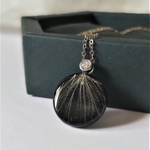Puede incluir: Un collar de cadena de plata con un colgante redondo negro que presenta un delicado dise&ntilde;o de diente de le&oacute;n blanco y un peque&ntilde;o acento de cristal transparente.